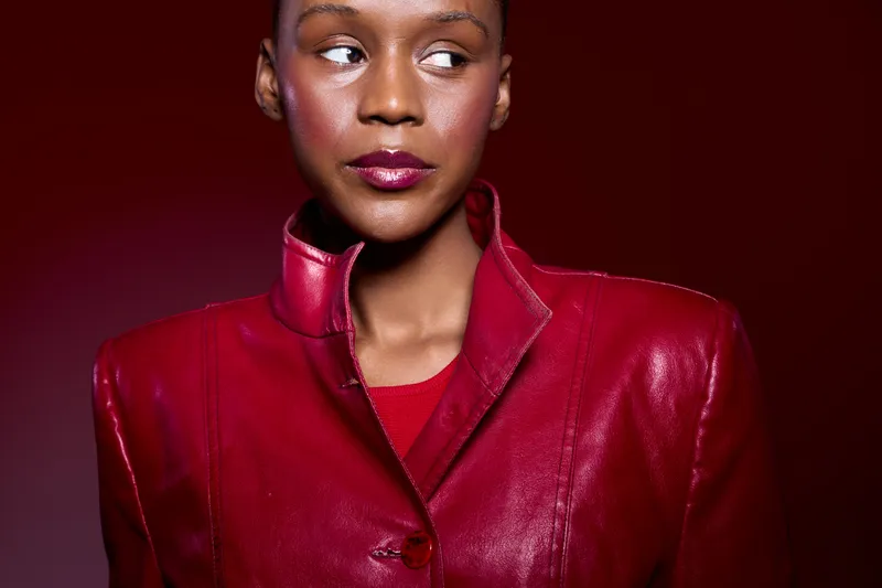 Portrait of confident black woman with dark skin wearing red leather jacket against dark red background. Concept of fashion, elegance, and modern style for female empowerment.