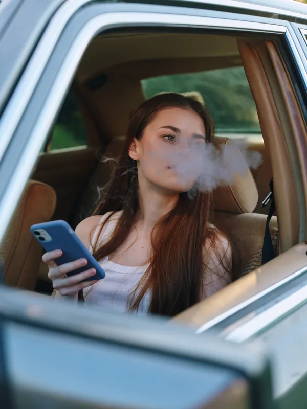 Young woman with long hair in casual attire smoking while relaxing in the car, surrounded by nature. Lifestyle, relaxation, and freedom are depicted here.