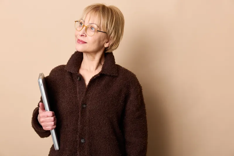 Senior woman with glasses holding a smartphone, thinking expression, calm profile, beige background, concept technology use for communication portrait