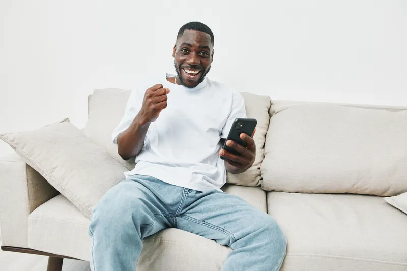 Joyful Black man celebrating success while holding a smartphone on a cozy couch, showcasing emotions of happiness and relaxation