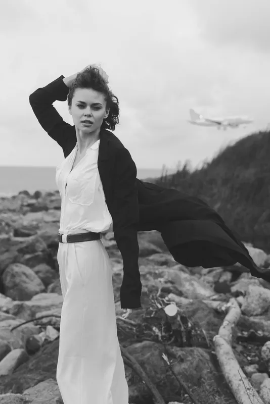 Woman on a windy rocky coast in a flowing dress and cardigan, dramatic black and white scene with a distant airplane, conveying fashion and rugged seascape for editorial use and mood