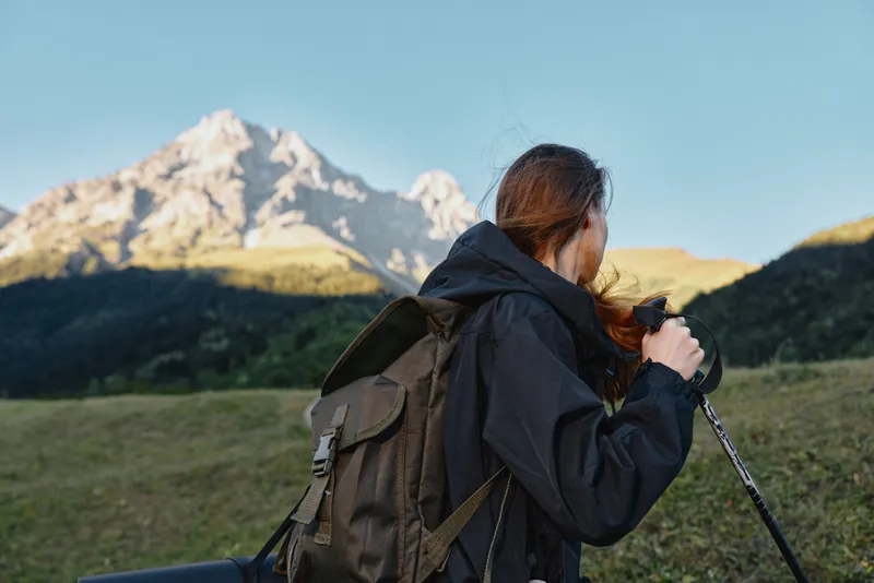 A photographer with a backpack stands in a mountain landscape, holding a camera and looking toward distant peaks, capturing a serene outdoor scene.