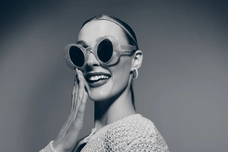 Smiling white woman with pale skin wearing large round sunglasses and earrings touches her face. Studio portrait in bright lighting, fashion concept with joyful mood and trendy accessories.