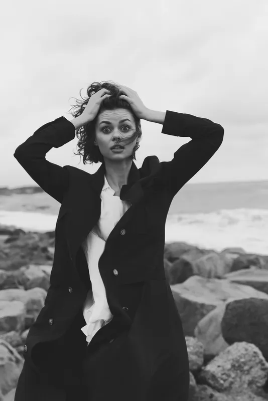 Woman on a rocky coastline in a long coat, wind swept, hands in hair, black and white mood, ocean backdrop, candid fashion editorial for dramatic seaside style and contrast