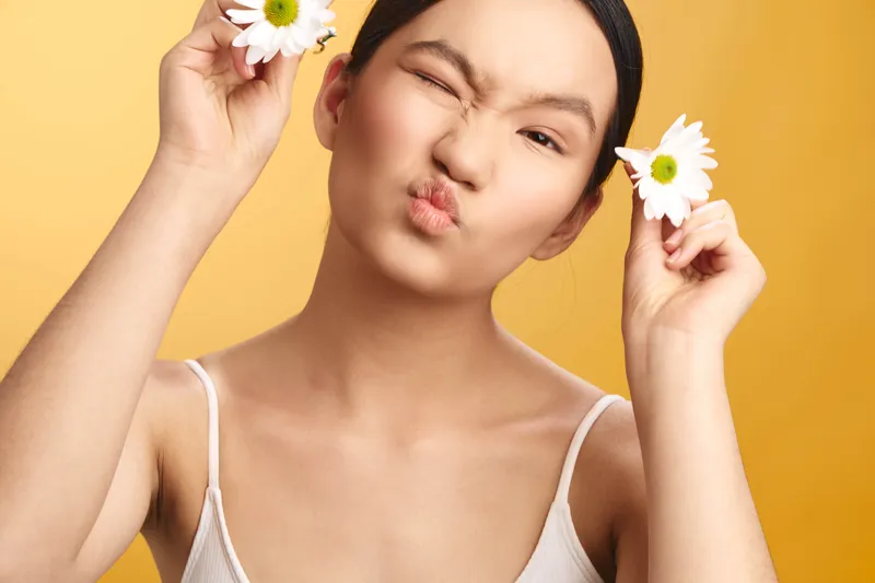 Asian woman smiles with flowers held beside her head in a playful pose against a bright backdrop, exuding charm and fresh beauty in a cheerful studio shot.