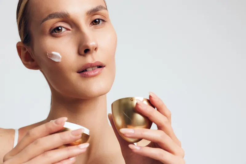 Young woman applying face cream with soft natural light, close-up portrait, clear skin, skincare routine, minimal makeup, neutral background, beauty concept