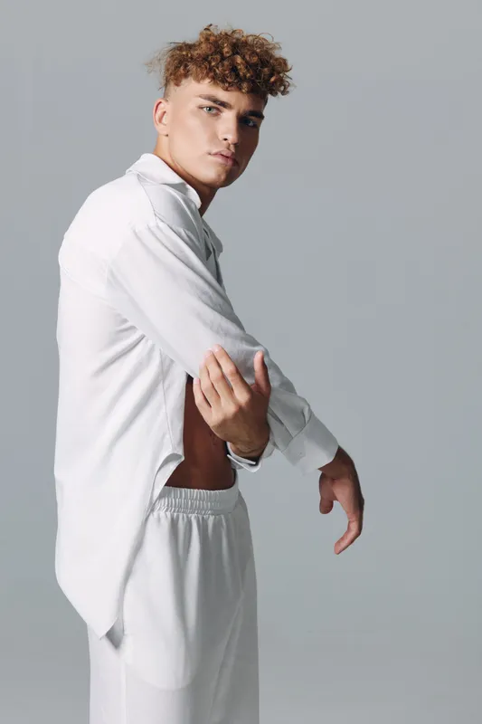 Young man with curly hair wearing white shirt and pants posing in studio with serious expression and neutral background for fashion or lifestyle concept.