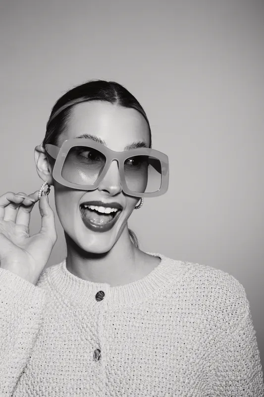 Young caucasian woman with light skin wearing large sunglasses and knitted sweater smiles joyfully on grey background studio.