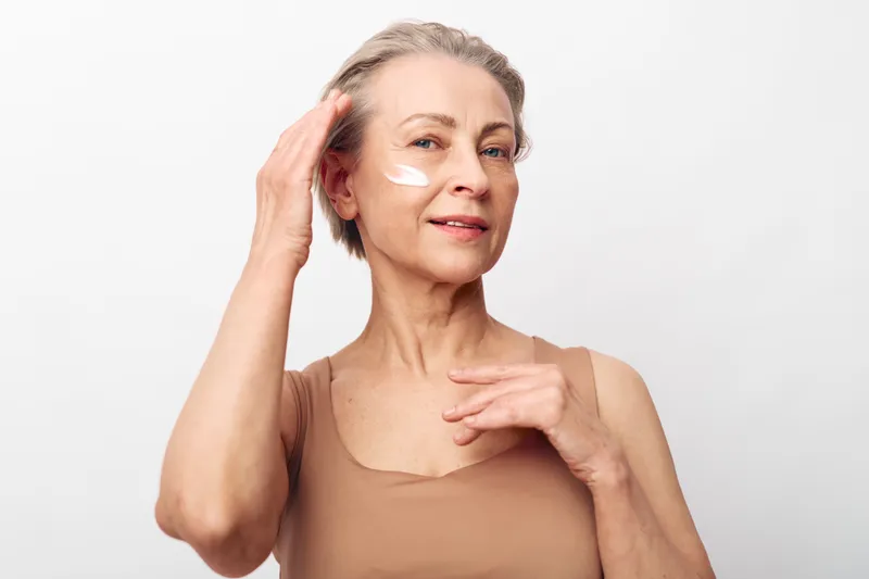 Mature woman with short hair applies moisturizer cream on her face, smiling softly. Skincare and self-care concept with natural beauty and healthy skin in bright studio setting.