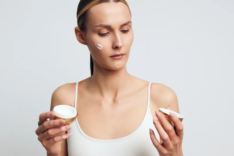 Young woman in white tank top applies face cream on cheek holds jar of moisturizer skincare product with focused expression isolated on light gray background.