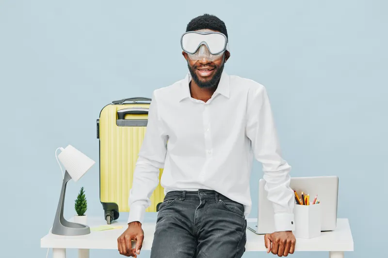 Smiling man wearing protective goggles and white shirt, standing at modern desk with travel essentials like yellow suitcase, white lamp, and stationery, inside a bright studio. Lifestyle concept.