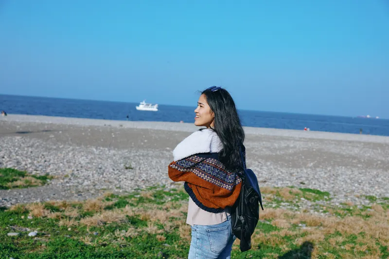 Woman with backpack on a sandy beach, looking toward the sea, wearing a scarf, sweater and jeans, clear sky and calm coast, outdoor travel mood and relaxation
