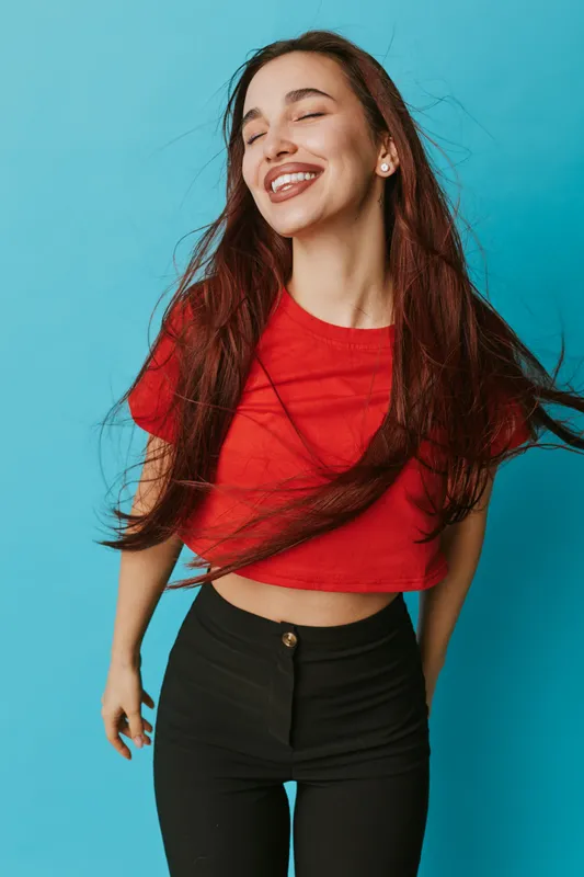 Young woman with long hair wearing red crop top and black pants smiling with eyes closed on blue background. Portrait of happy female model enjoying moment with carefree expression.