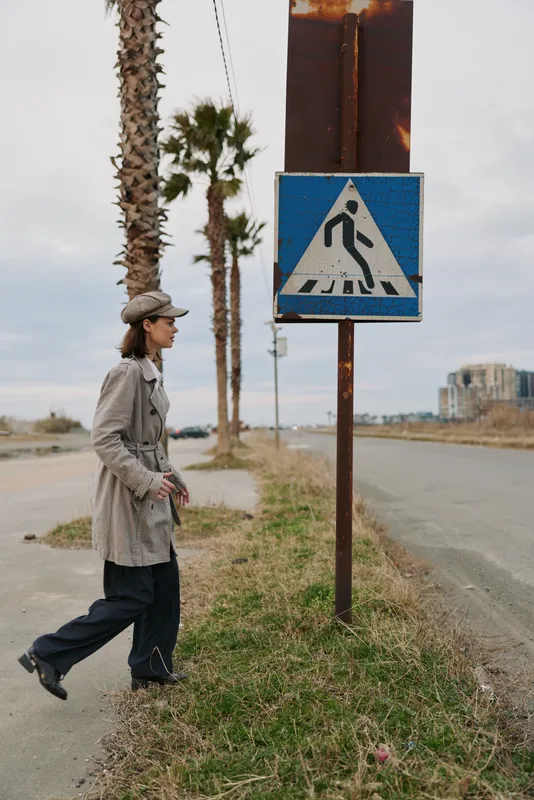 A woman in a trench coat and hat walks on the roadside near a pedestrian crossing sign, palm trees in the background on a cloudy day. Urban lifestyle concept.