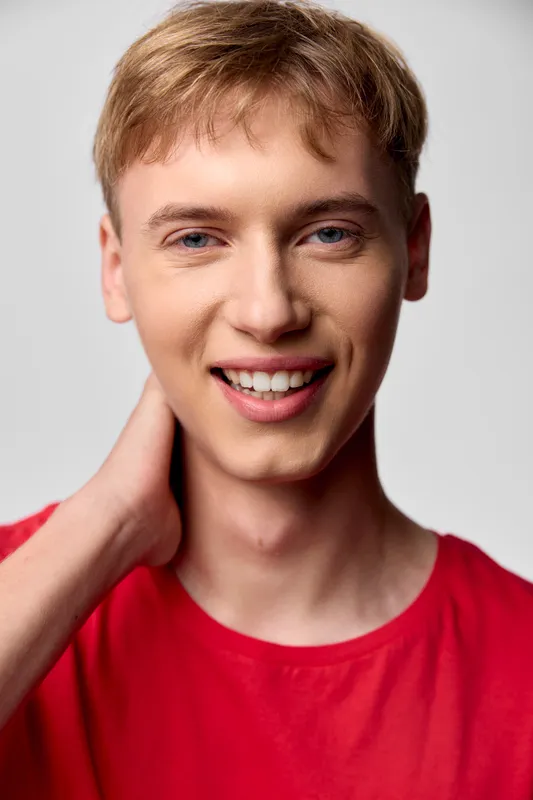 Smiling young man in a red shirt, hand at neck, soft studio lighting and approachable vibe for lifestyle, fashion, and marketing stock imagery.