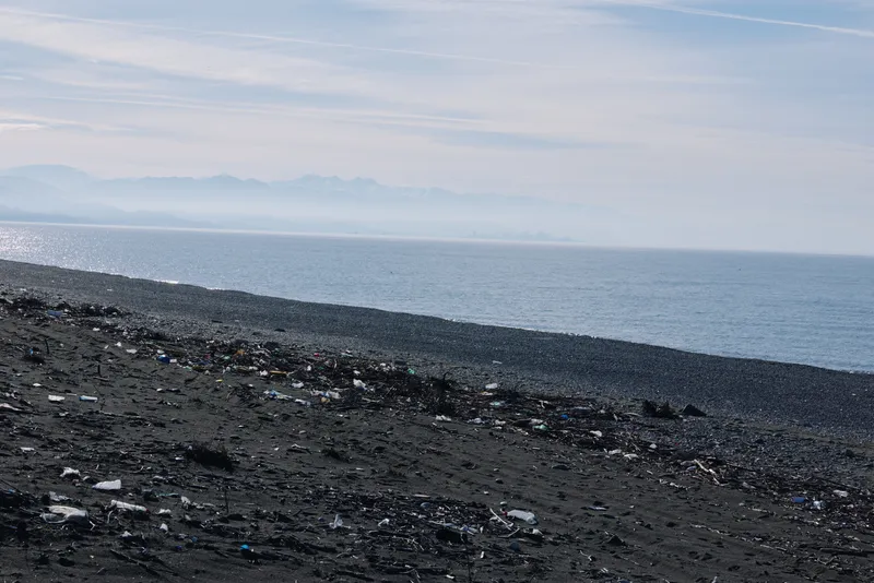 Dark pebble beach meets calm sea and distant hazy mountains on the horizon, a minimalist seascape highlighting coastline, water and sky in tranquil tones, with an empty shoreline inviting reflection