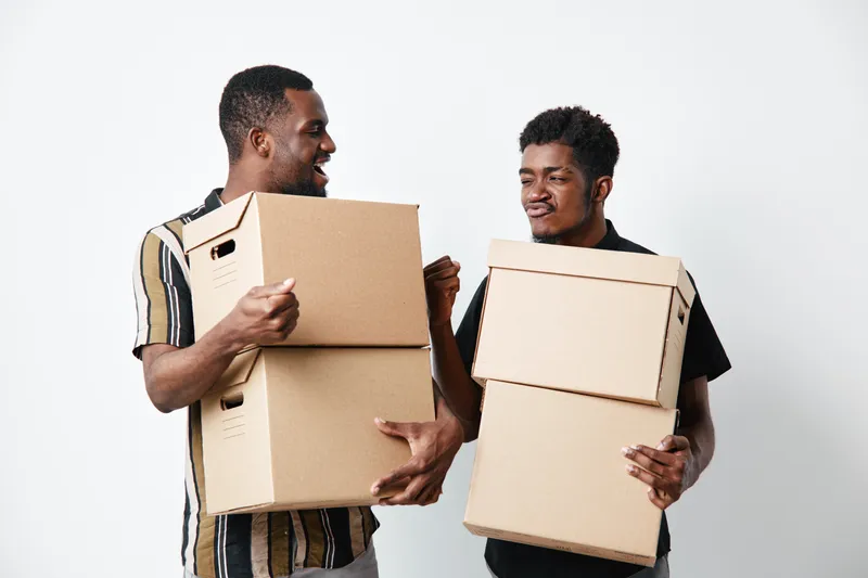 Two black men with dark skin carrying cardboard boxes and smiling while moving or working together in casual clothes on white background for teamwork and delivery.