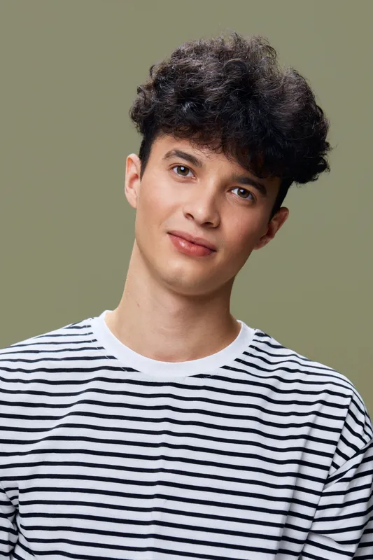 young man wearing white and black striped shirt smiling softly with curly dark hair and neutral green background, casual and friendly portrait