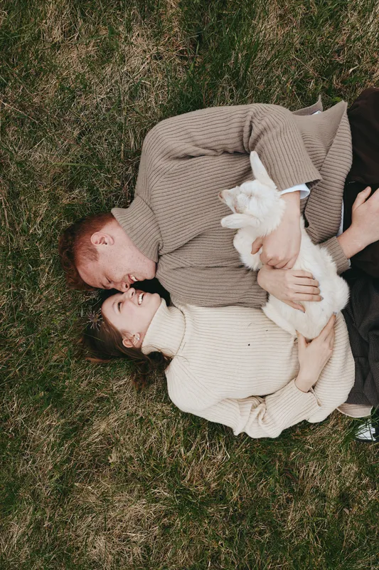 Candid couple outdoors, embracing on grass with a playful goat kid nestled between, warm sweater tones, tender smiles, relaxed mood, natural light, intimate moment, connection with nature, lifestyle