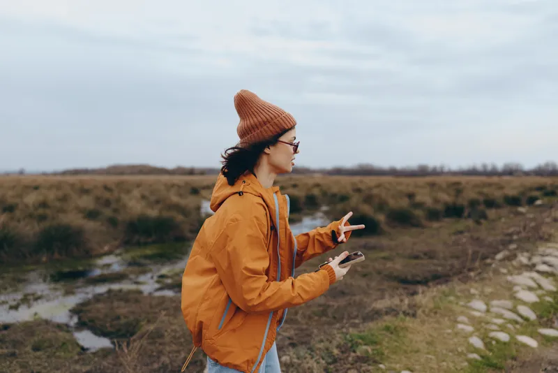 Woman outdoor lifestyle portrait autumn countryside, wearing orange jacket and knit hat, walking along rural path and holding phone, casual travel mood, natural daylight