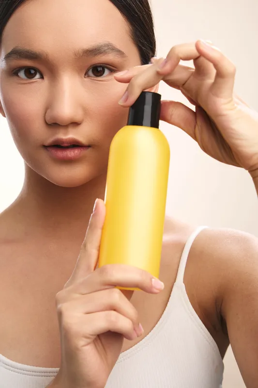 Asian woman holds a bright yellow bottle in a close up shot, showcasing a skincare product. She looks calm and focused, creating a clean, modern beauty moment.
