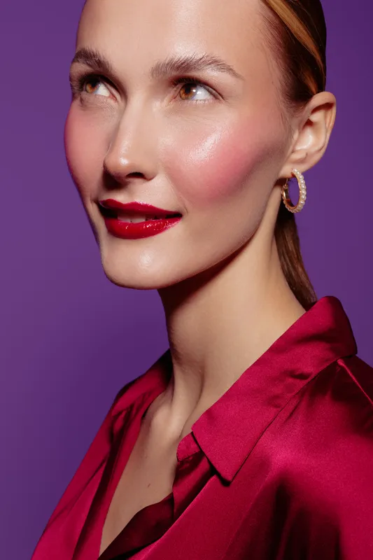 Close-up portrait of a woman with glossy red lips and pink blush wearing a red satin blouse and gold hoop earrings on purple background. Concept of beauty, makeup, and fashion.