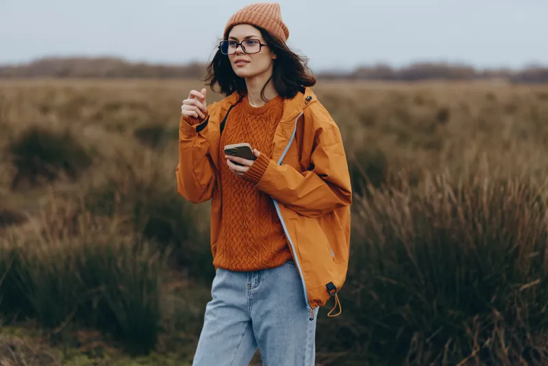 Woman lifestyle portrait outdoors in autumn field, wearing beanie and glasses, orange jacket over sweater, holding coffee cup, walking through grassy landscape, relaxed mood, natural light