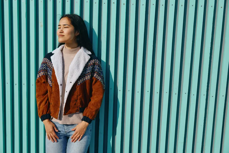 Fashion, style, outfit, cardigan, jeans, urban elements describe a woman standing beside a teal striped wall, wearing a cozy patterned cardigan and light sweater for a relaxed, confident urban vibe.