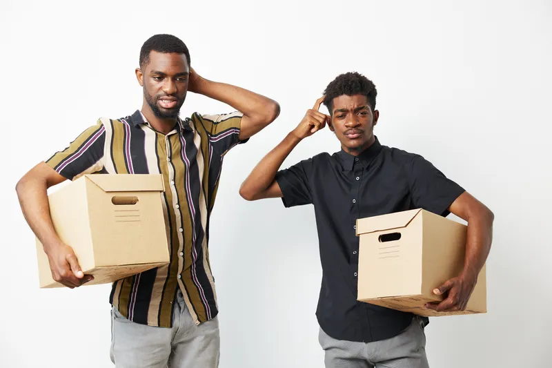 Two black men standing with cardboard boxes, looking confused and scratching their heads, showcasing expressions of uncertainty and contemplation.