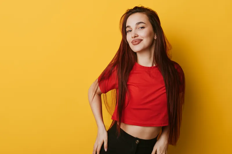 Young woman with long hair and red shirt posing confidently against yellow background. Casual style, smiling face and relaxed posture for lifestyle and fashion portrait.
