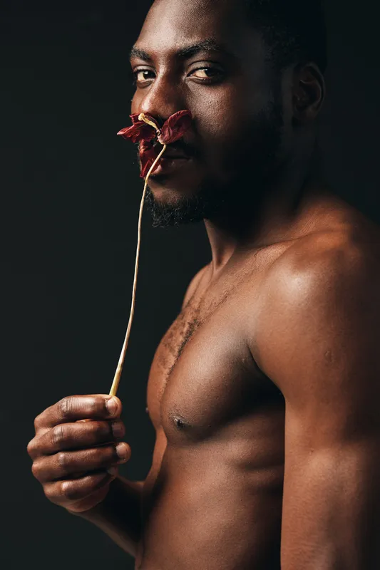 African man with dark skin holding dried flower near nose in creative conceptual visual style. Portrait, bare chest and artistic expression with moody lighting on black background.