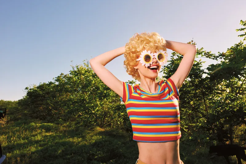 Smiling woman with curly blonde wig wearing a striped cropped top outdoors in bright sun. Playful retro style, outdoor lifestyle, and carefree mood with vibrant colors.