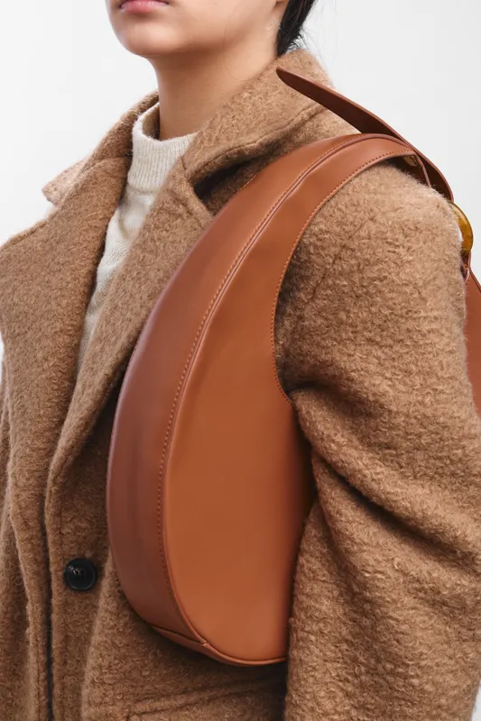 Asian woman wearing a warm brown coat and a sleek leather bag sits in a modern setting, projecting confidence, style, and a professional, approachable vibe.