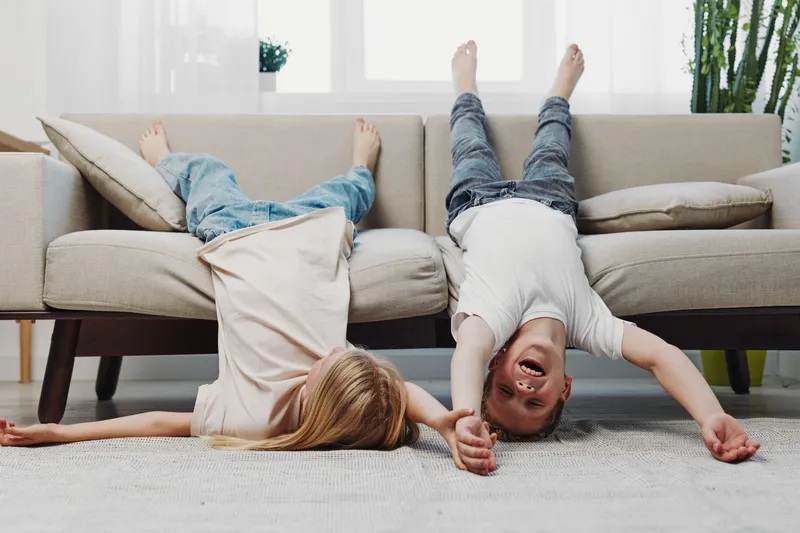 Two happy children lie upside down on a sofa in a bright living room, laughing and holding hands. Concept of childhood joy, playfulness, and carefree moments indoors.