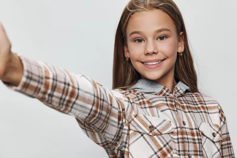 Girl with braces smiling and taking selfie with outstretched arm wearing plaid shirt isolated on white background. Child, happiness and casual clothing in studio.