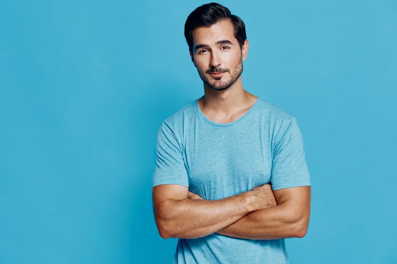 Handsome young man wearing a light blue t-shirt standing confidently with arms crossed against a solid blue background. Casual lifestyle concept.
