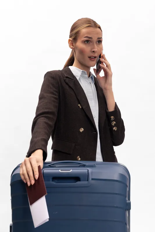 Concerned businesswoman in formal jacket holds passport and boarding pass, standing with large suitcase while talking on phone. Concept of travel, business trip, and urgent communication.