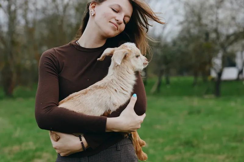 Joyful woman outdoors holding a goat kid in her arms, park setting, casual sweater, candid lifestyle moment, natural light, warm mood, gentle bond with animal, authentic and serene countryside scene
