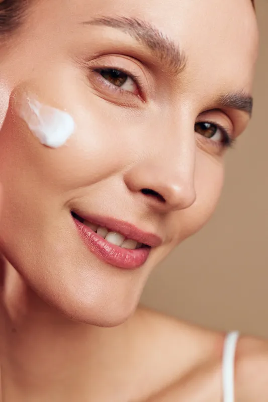 Close-up of smiling woman applying white face cream on cheek. Natural skin texture visible, soft lighting highlights healthy complexion. Concept of skincare and beauty care.