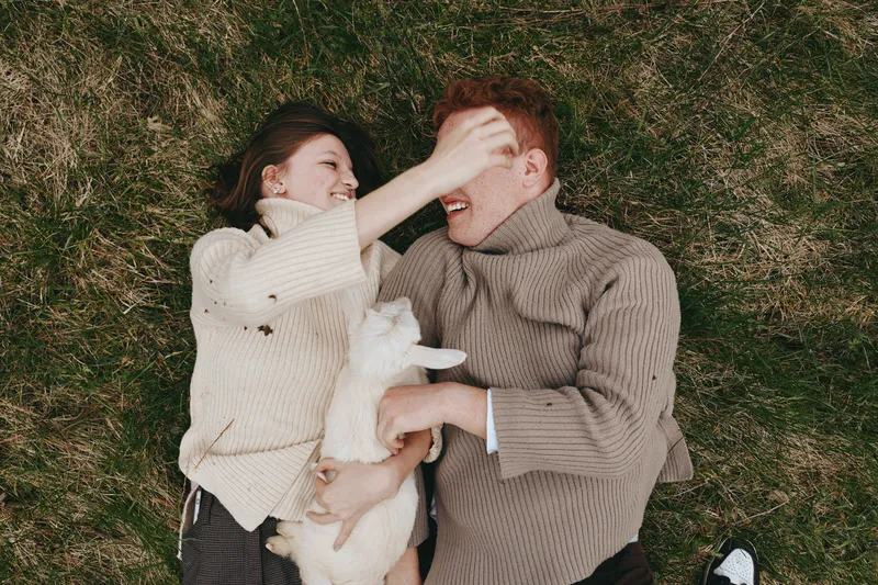 Couple outdoor grass goat kid cuddle affection cozy sweater lifestyle romance joyful smile in nature sunlight on meadow field warmth bond laughter peaceful moment