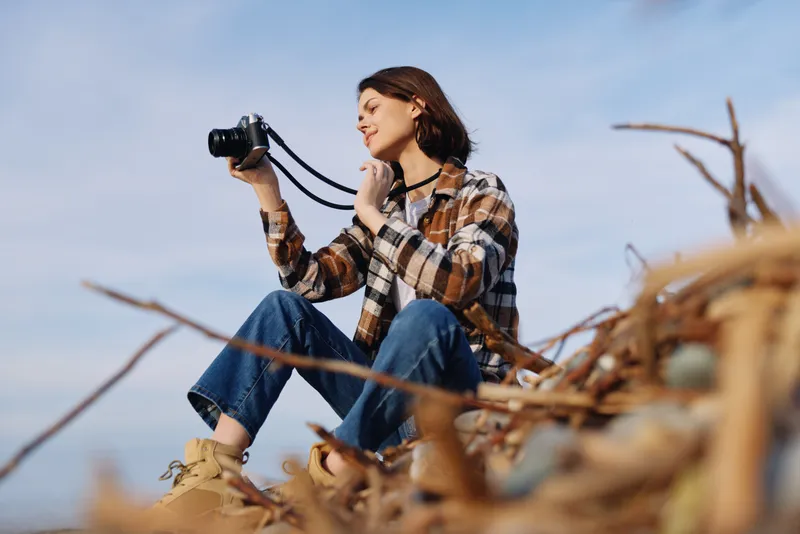 Woman photographer sitting on scattered branches outdoors, holding a camera and looking through the viewfinder, embracing nature and a calm, creative moment in a sunny open setting