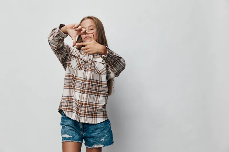 Girl in plaid shirt and denim shorts posing with hands framing face on light background for childhood, happiness, casual style, and playful mood photo.
