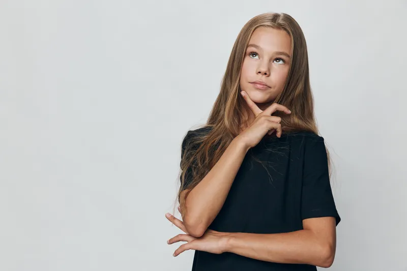 Girl teenager with long hair in black shirt thinking and looking up on gray background. Child expression with hand on chin for concentration and decision making.