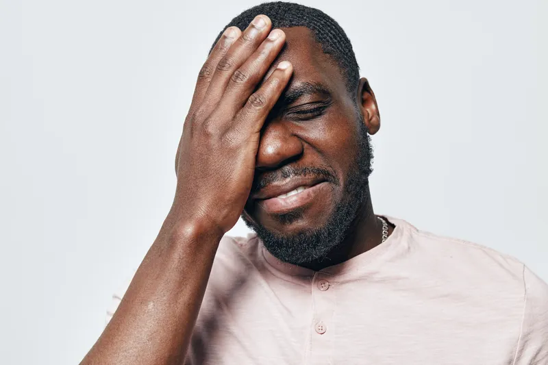 African American man with dark skin and beard wearing light shirt touching face with hand and eyes closed in emotional expression on white background.