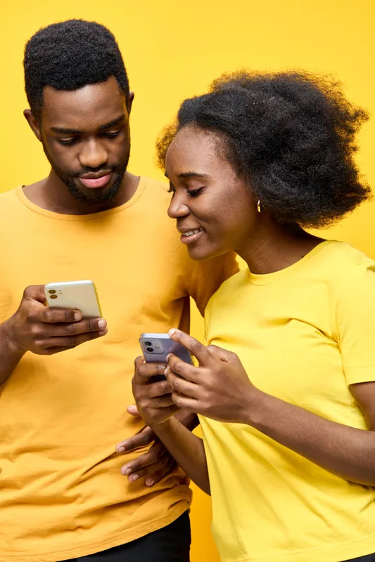 Young couple sharing smartphones with joyful expressions against a vibrant yellow background, embodying modern communication and connection in a cheerful setting
