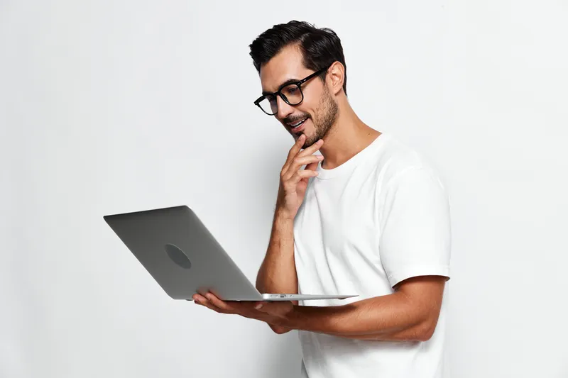 Smiling man with glasses holding laptop, looking thoughtfully at screen, isolated on plain white background. Technology and lifestyle concept.