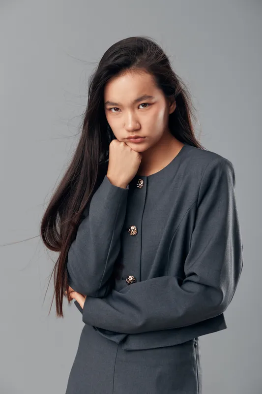Young woman with long dark hair posing with serious expression and folded arms against plain gray background in studio, wearing gray long-sleeve top with buttons.