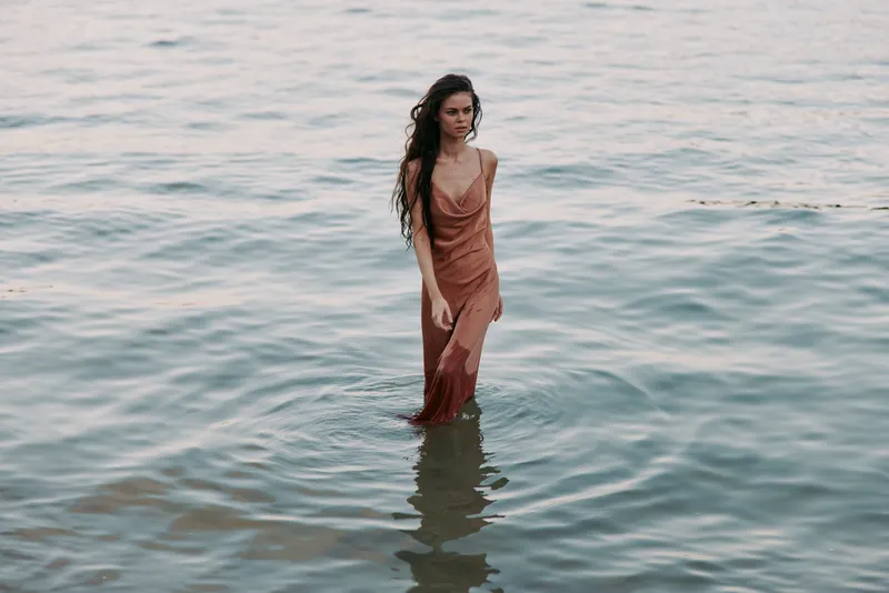 A solitary woman stands in shallow sea water wearing a flowing long dress; gentle waves lap at her legs, creating a serene, sunlit coastal moment of calm, quiet beauty.