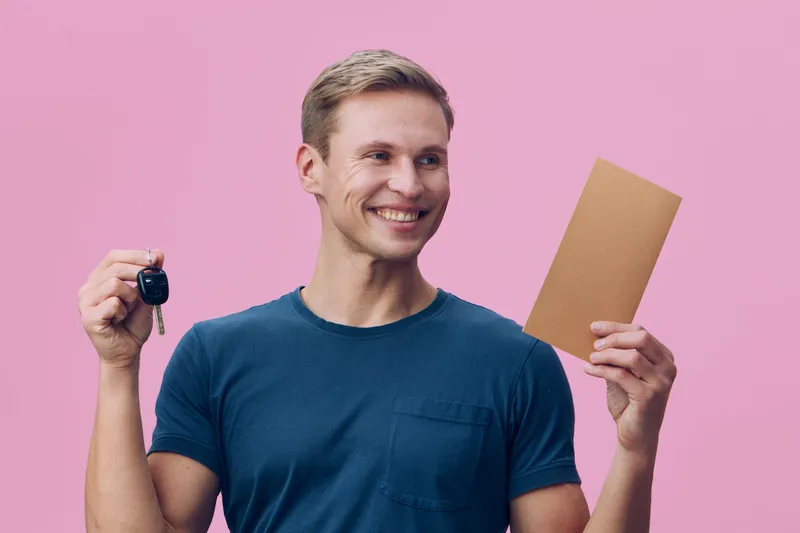 Smiling young man holding a car key in one hand and a cardboard box in the other, standing against a pink background. Casual clothing, cheerful expression, victory gesture. Studio shot, lifestyle conc