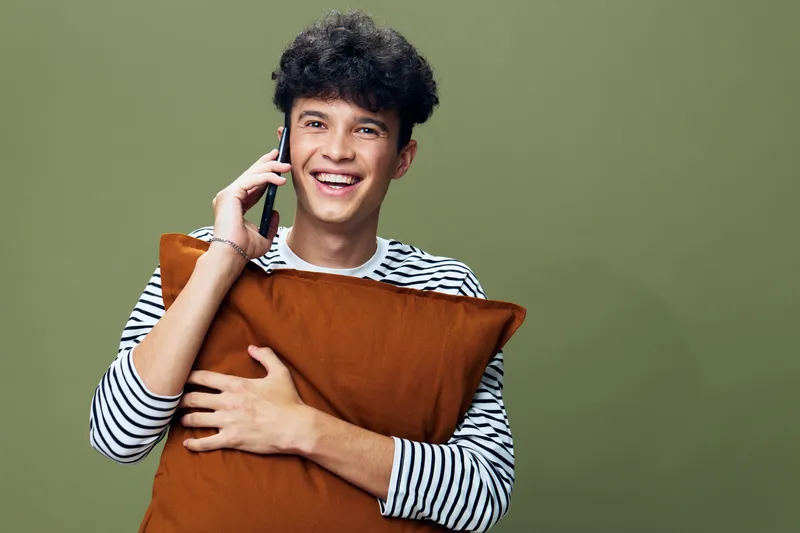 happy young man smiling while talking on phone and holding brown pillow, casual clothing, green background, cheerful expression, positive mood